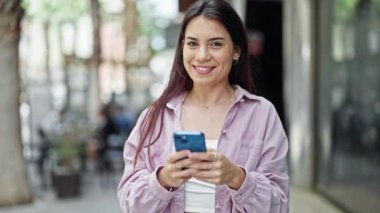 Genç, güzel, İspanyol bir kadın sokakta akıllı telefon kullanarak gülümsüyor.