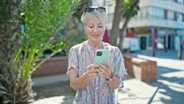 中年ブロンドの女性観光客が路上でスマートフォンを使用して自信を持って微笑む — ストック動画