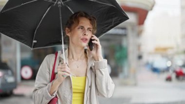 Şemsiye tutan genç bir kadın akıllı telefondan konuşuyor. Sokakta gülümsüyor.