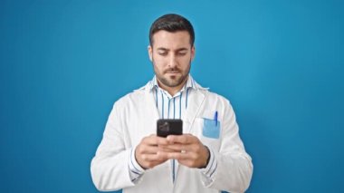 Genç İspanyol doktor mavi arka planda akıllı telefon kullanarak gülümsüyor.