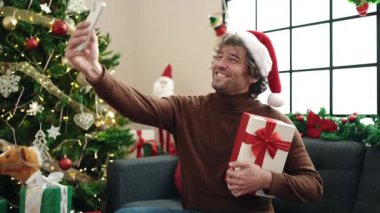 Genç İspanyol adam akıllı telefon kullanıyor. Kanepede oturuyor. Elinde Noel hediyesi var.