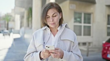 Orta yaşlı İspanyol kadın sokakta akıllı telefon kullanıyor.