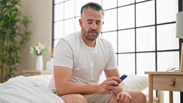 头发灰白的男人坐在床上用智能手机看着卧室很不舒服 — 图库视频影像