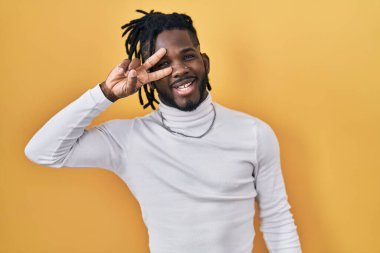 Sarı arka planda balıkçı yaka kazak giyen Afrikalı bir adam barış sembolü yapıyor. Yüzünde parmakları var, gülümsüyor ve zaferini gösteriyor. 