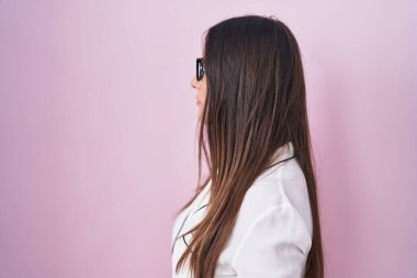 Genç esmer kadın pembe arka planda duran gözlüklü, yan yan yan bakan, rahat poz veren, kendine güvenen gülümsemeli.. 