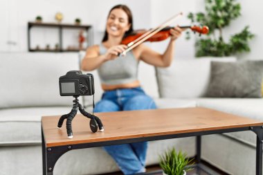 Young beautiful hispanic woman recording violin class sitting on sofa at home