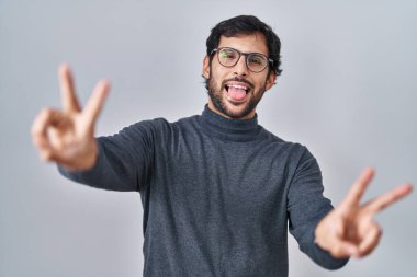 İzole edilmiş arka planda duran yakışıklı Latin adam iki elini de zafer işareti yaparak parmaklarını göstererek gülümsüyor. İki numara.. 