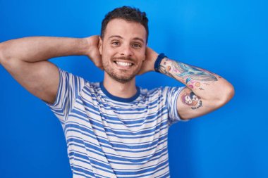Young hispanic man standing over blue background relaxing and stretching, arms and hands behind head and neck smiling happy 