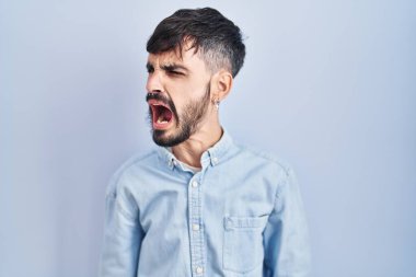 Young hispanic man with beard standing over blue background angry and mad screaming frustrated and furious, shouting with anger. rage and aggressive concept. 