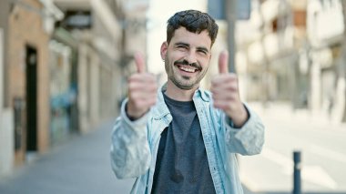Young hispanic man smiling confident doing ok sign with thumbs up at street