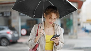 Sokakta gülümseyen akıllı telefon kullanan şemsiyeli genç bir kadın.