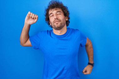 İspanyol genç adam mavi arka planda dikiliyor, geriniyor, yorgun ve rahat, uykulu ve sabahın erken saatleri için esniyor. 