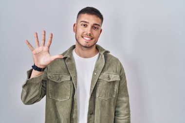 İzole edilmiş arka planda duran genç İspanyol adam beş numarayı gösterip işaret ederken kendinden emin ve mutlu bir şekilde gülümsüyor.. 