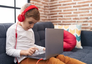 Adorable hispanic boy using laptop and headphones sitting on sofa at home