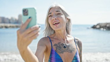 Orta yaşlı, kır saçlı, bikini giyen turist kumsalda video görüşmesi yapıyor.