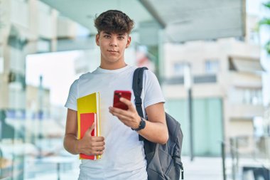 Young hispanic teenager student using smartphone holding books at university
