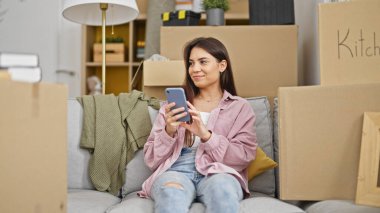 Genç ve güzel İspanyol kadın akıllı telefon kullanıyor. Yeni evindeki koltukta oturuyor.