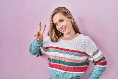 Young blonde woman standing over pink background smiling looking to the camera showing fingers doing victory sign. number two. 