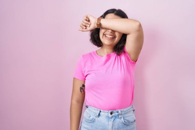 Pembe arka planda duran genç İspanyol kadın kollarıyla gözlerini kapatıyor neşeli ve komik gülümsüyor. kör kavram. 