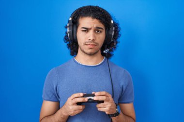 Hispanic man with curly hair playing video game holding controller skeptic and nervous, frowning upset because of problem. negative person. 