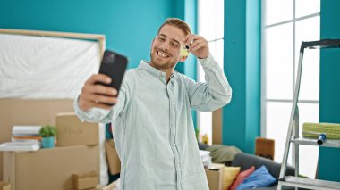 Kafkasyalı genç adam yeni evinde elinde anahtarla akıllı telefondan selfie çekiyor.