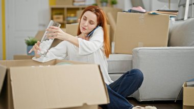 Karton kutuları açan genç kızıl saçlı kadın yeni evinde telefonla konuşuyor.