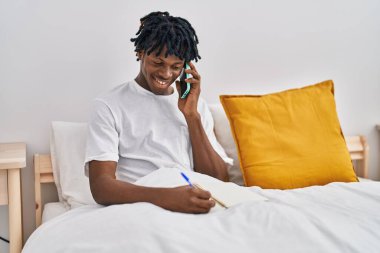 African american man talking on the smartphone writing on notebook sitting on bed at bedroom