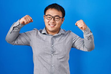 Mavi arka planda duran genç Çinli adam gururla gülümseyen kol kaslarını gösteriyor. fitness konsepti. 