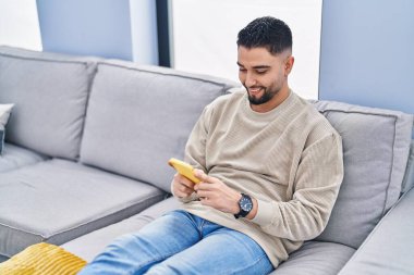 Young arab man using smartphone sitting on sofa at home
