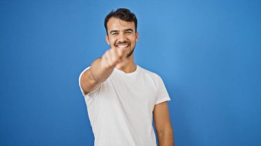 Genç İspanyol adam gülümsüyor, kendine güveni tam, mavi arka planda kamerayı işaret ediyor.
