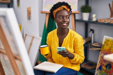 Genç Afrikalı Amerikalı kadın sanatçı akıllı telefon kullanıyor. Sanat stüdyosunda kahve içiyor.