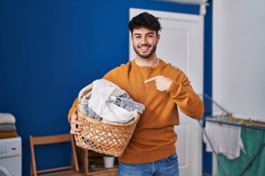Çamaşır odasında çamaşır sepetini tutan sakallı İspanyol bir adam mutlu ve gururlu bir kendini işaret ediyor. 