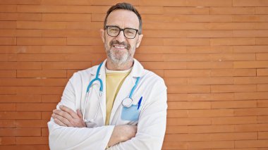 Orta yaşlı doktor güler yüzlü. Tuğladan yapılmış arka planda duruyor.