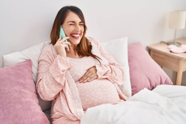 Genç hamile bir kadın akıllı telefonda konuşuyor. Yatak odasında oturuyor.