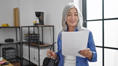 Ofiste evrak okuyan orta yaşlı, kır saçlı bir iş kadını.