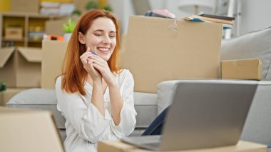 Genç kızıl saçlı kadın yeni evinde laptop ve kredi kartıyla alışveriş yapıyor.