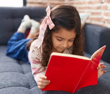 Adorable hispanic girl reading book lying on sofa at home