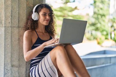 Young beautiful hispanic woman listening to music sitting on bench at street