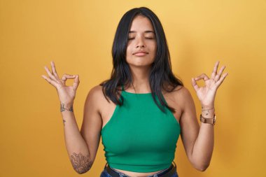 Esmer kadın sarı arka planda duruyor rahatlamış ve gözleri kapalı gülümsüyor parmaklarıyla meditasyon hareketi yapıyor. yoga konsepti. 