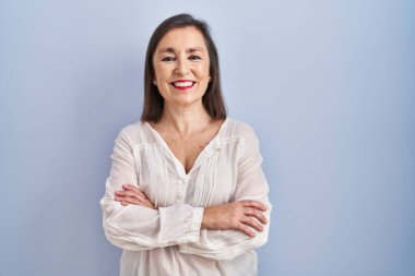 Orta yaşlı İspanyol kadın mavi arka planda durup kollarını kavuşturup kameraya bakarken gülümsüyor. pozitif kişi. 