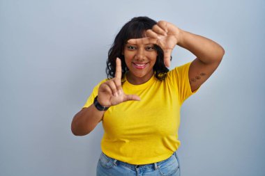 Hispanic woman standing over blue background smiling making frame with hands and fingers with happy face. creativity and photography concept. 