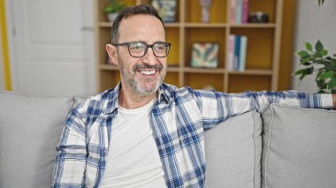 Orta yaşlı bir adam evdeki koltukta otururken kendinden emin gülümsüyor.