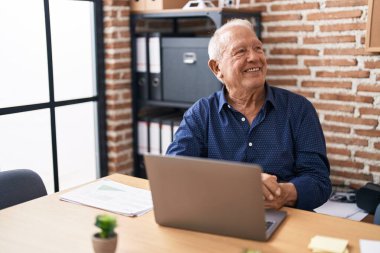 Ofiste bilgisayar bilgisayarı kullanarak çalışan yaşlı adam yüzünde gülümseme ve doğal ifadeyle yan yana bakıyor. Kendinden emin gülen. 