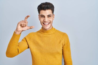 Mavi arka planda duran genç İspanyol adam gülümsüyor ve el hareketleriyle hareket ediyor parmaklarıyla işaret yapıyor ve kameraya bakıyor. ölçüm kavramı. 