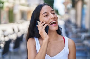 Genç, güzel, İspanyol bir kadın gülümsüyor ve caddede akıllı telefondan konuşuyor.