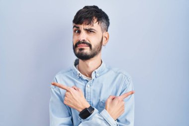 Mavi arka planda duran sakallı genç İspanyol adam iki tarafı da parmaklarıyla işaret ediyor, farklı yönlere. 
