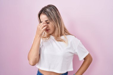 Pembe arka planda duran genç sarışın kadın yorgun burnu ve gözleri ovuşturup yorgun ve baş ağrısı çekiyor. Stres ve hüsran kavramı. 