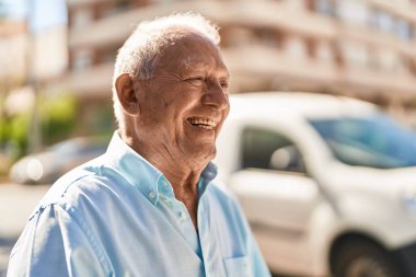 Yaşlı, kır saçlı, gülümseyen, kendine güvenen bir adam sokakta duruyor.