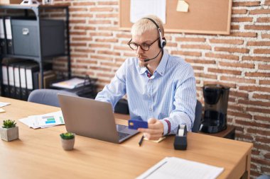 Young caucasian man call center agent using computer holding credit card at office