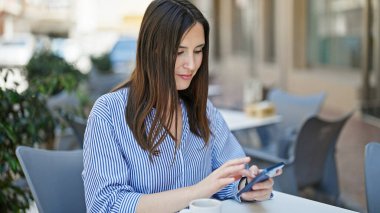 Akıllı telefon kullanan genç ve güzel bir İspanyol kadın kahve dükkanının terasında oturmuş gülümsüyor.